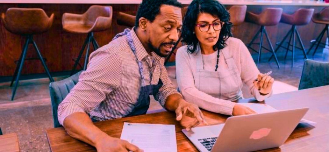 A Black man and an Indian woman wearing cafe aprons and sitting at a table in a cafe in front of a laptop conducting business